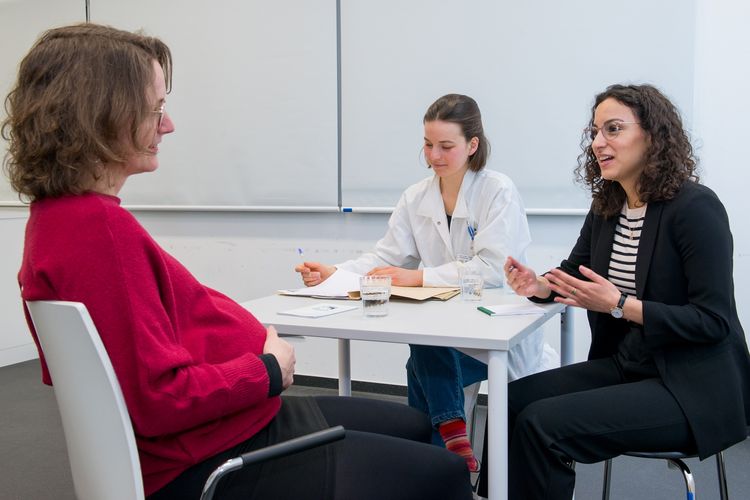 Drei Frauen sitzen um einen kleinen Tisch herum. Eine Frau ist schwanger und hört der Frau zu, die gerade mit ihr spricht. Eine weitere junge Frau trägt einen weißen Kittel und hat eine Akte aufgeschlagen vor sich liegen. Foto: Bodo Tiedemann