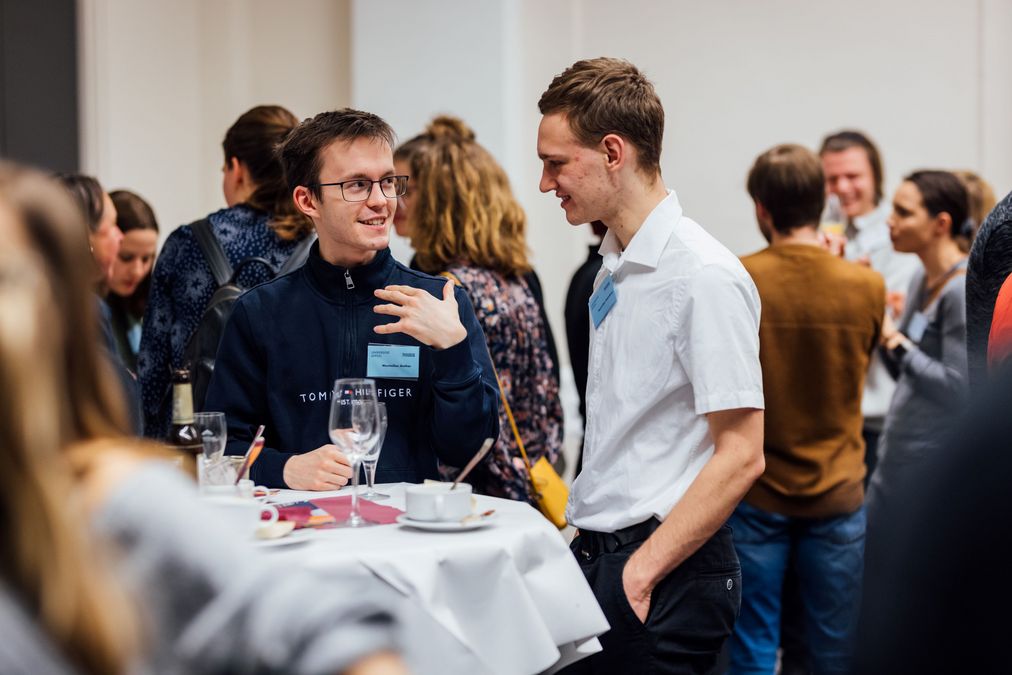 zur Vergrößerungsansicht des Bildes: Stipendiaten stehen an einem Stehtisch mit Speisen und Getränken und unterhalten sich