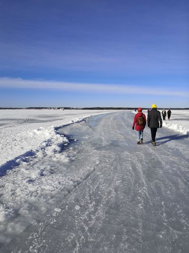 Zwei Drittel des Bildes werden durch eine zugeschneite, vereiste Fläche gefüllt. Zwei Personen laufen einen vereisten Weg entlang. Am Horizont sind ein paar Häuser zu sehen. Darüber befindet sich blauer Himmel.
