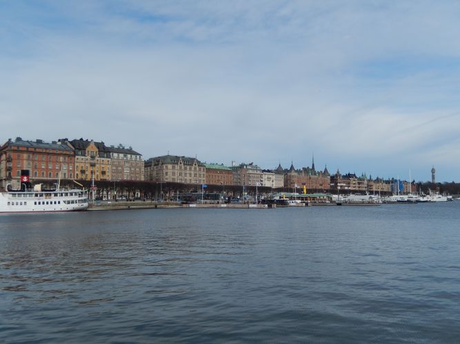 Hier sieht man einen Hafen und im Hintergrund eine boulevardähnliche Prachtstraße in Stockholm.
