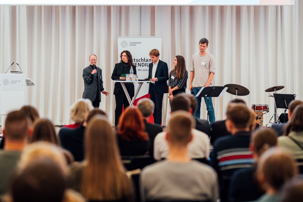 zur Vergrößerungsansicht des Bildes: auf der Bühne stehen die Moderatoren von mephisto 97.6 Clara Haferkamp und Leonard Roemer, Stipendiatin Maria Bönsch, Förderer: Optimax Energy GmbH mit Albert Mantel und Tina Steinbiß und sprechen zu den Gästen