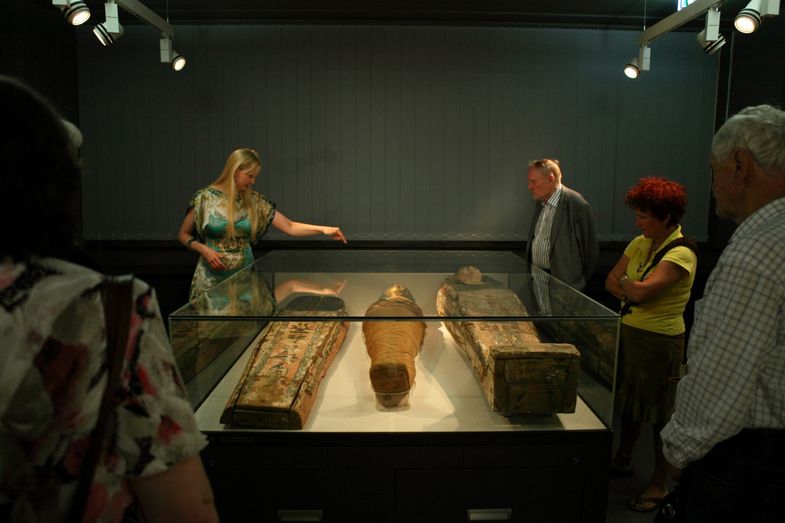 Blick in einen Raum mit einer Vitrine mit Mumien und Sarg, daneben Besucher und eine Mitarbeiterin