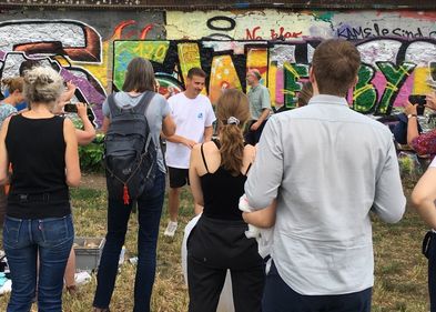 vor einer mit Graffiti besprühten Wand stehen junge Leute und hören einem Mann zu, der etwas erklärt