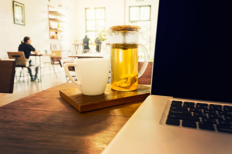 Auf einem Tisch steht ein Laptop, daneben steht eine Tasse und eine Glaskanne mit Tee. Im Hintergrund ist eine Szene im Café mit einer Person an einem Tisch sitzend zu erkennen.