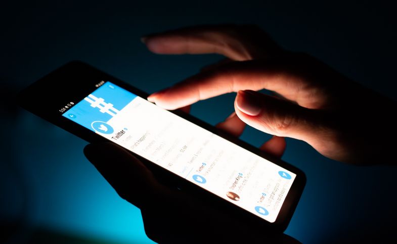 Symbolbil: Dunkler Hintergrund, der Bildschirm eines Smartphones leuchtet es ist der Schriftzug Twitter zu sehen. Eine Hand tippt auf das Smartphone.