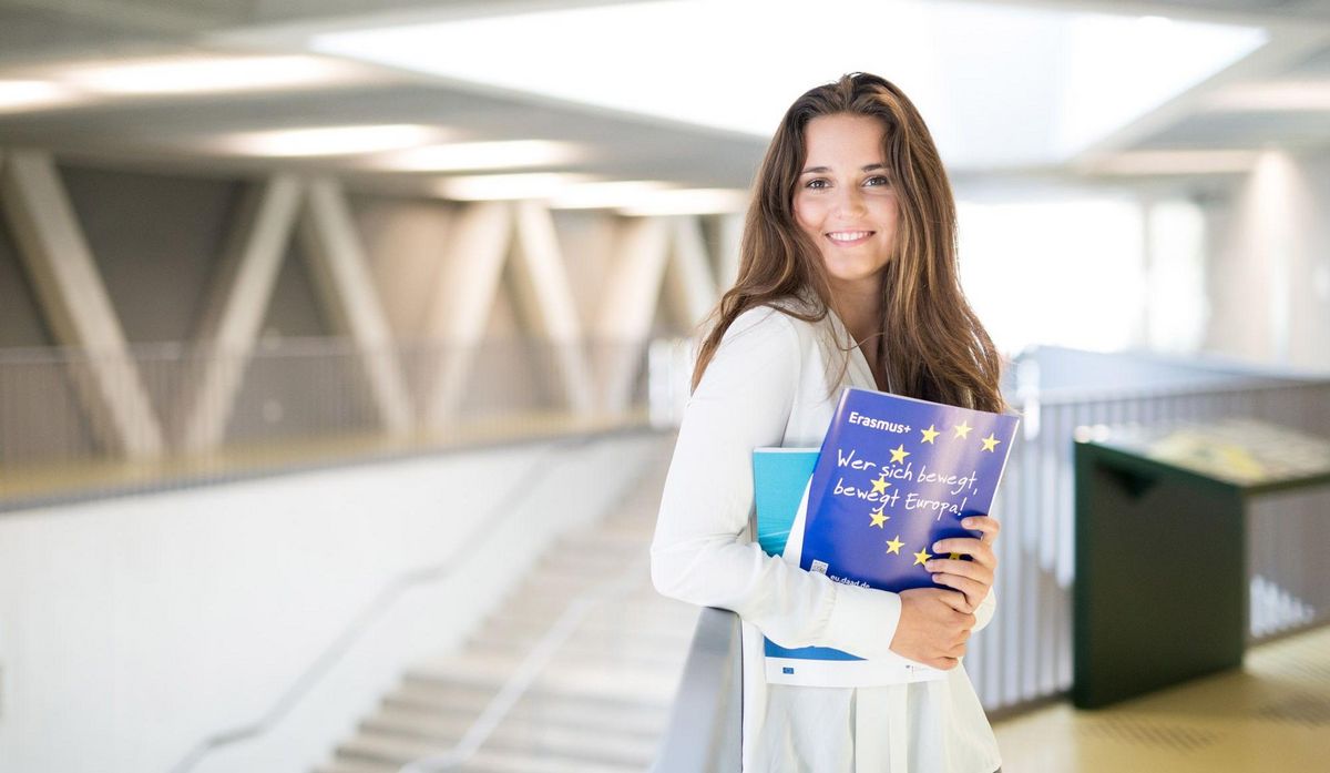 zur Vergrößerungsansicht des Bildes: Junge Frau mit Erasmus Prospekten steht in der Uni