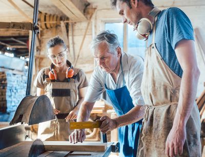 Man sieht drei Personen aus verschiedenen Generationen in einer Tischlerwerkstatt. Ein älterer Herr bringt einer jungen Frau und einem jungen Mann bei, mit einer Holzverarbeitungsmaschine zu arbeiten.