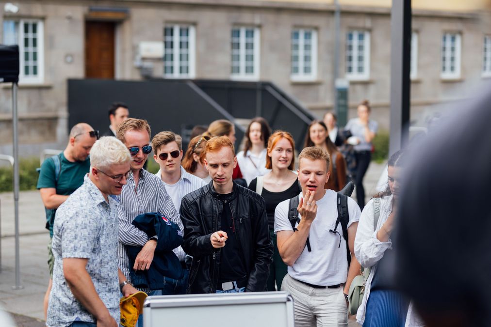 zur Vergrößerungsansicht des Bildes: vier Studenten stehen vor einem Aufsteller und informieren sich über die angebotenen Führungen, im Hintergrund sind weitere Studierende, die sich unterhalten und warten