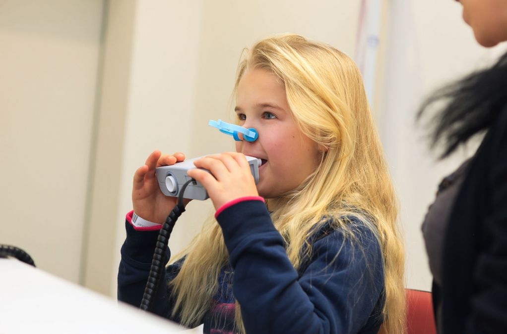 Ein Mädchen macht einen Lungenfunktionstest in der LIFE-Child-Studienambulanz. Sie hat eine Klemme auf der Nase und hät ein Messgerät in den Händen, in das sie hinein pustet.