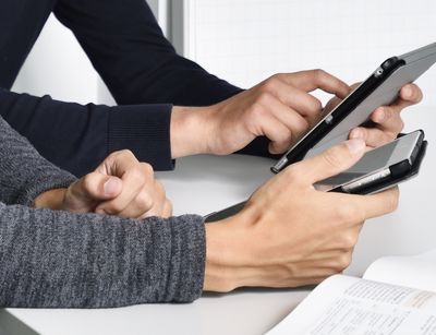 Zu sehen sind die Arme zweier Personen, die am Tisch sitzen und Tablets in der Hand halten. 