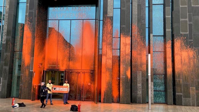 Zwei Protestierende stehen mit einem Transparent vor dem orange besprühten Augusteum mit einem Protestbanner in der Hand