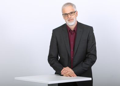 Ein Mann mit dunklem Anzug und rotem Hemd, grauem Vollbart und Brille steht an einem Tisch und schaut in die Kamera. Professor Dr. Gert Pickel, Foto: Universität Leipzig.