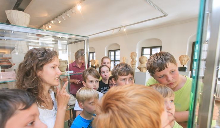 Eine Gruppe Kinder schaut interessiert in eine Vitrine des Antikenmuseums. Der Fokus liegt auf den Gesichtern der Kinder, die Exponte sind nicht abgebildet.