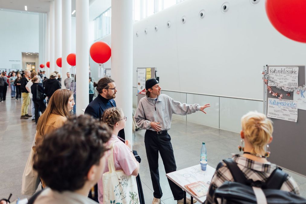 zur Vergrößerungsansicht des Bildes: Foto: Besucher:innen am Infostand im Gespräch mit einem Studenten.