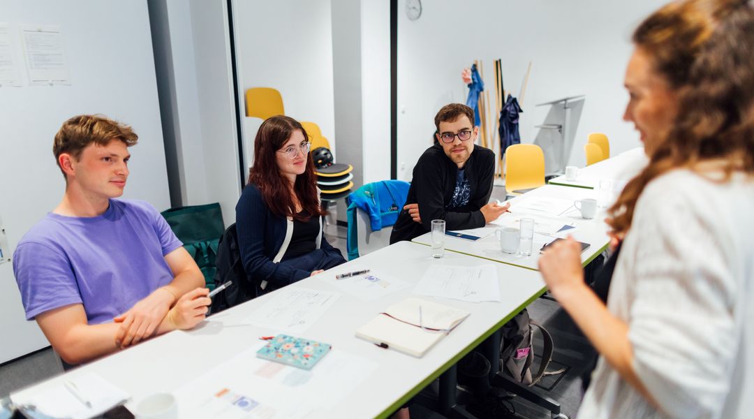 Farbige Aufnahme von drei Personen, die nebeneinander an einem Tisch sitzen und zu einer vor ihr stehenden Person blicken