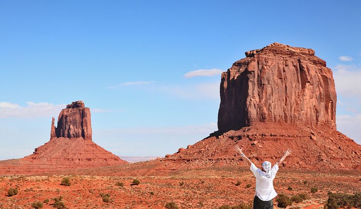 Mensch steht mit dem Rücken zur Kamera vor einer Wüstenlandschaft mit Bergen und streckt die Hände in die Höhe.