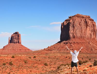 Person stands with their back to the camera in front of a desert landscape with mountains and stretches their hands up in the air.