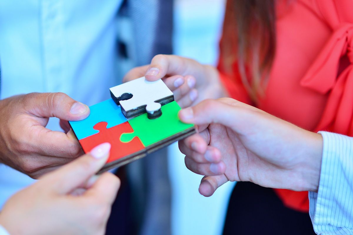 zur Vergrößerungsansicht des Bildes: Detailaufnahme: Vier Hände halten je ein Puzzleteil aus Holz. Das blaue, rote und grüne Puzzleteil stecken fest zusammen. Das weiße Puzzleteil ist in etwas erhöhter Position und wird gerade hinzugefügt. Alle vier Teile zusammen ergeben ein Quadrat.