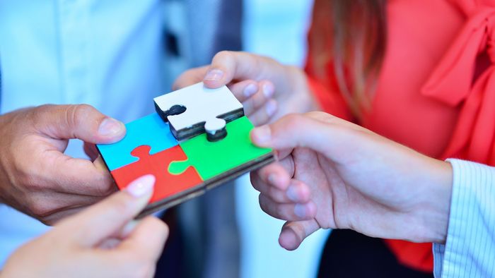 Detailaufnahme: Vier Hände halten je ein Puzzleteil aus Holz. Das blaue, rote und grüne Puzzleteil stecken fest zusammen. Das weiße Puzzleteil ist in etwas erhöhter Position und wird gerade hinzugefügt. Alle vier Teile zusammen ergeben ein Quadrat.