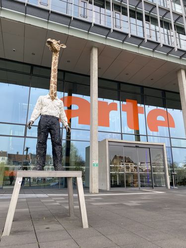 Bild einer Giraffenmannskulptur, die sich vor einem Arte-Gebäude befindet.