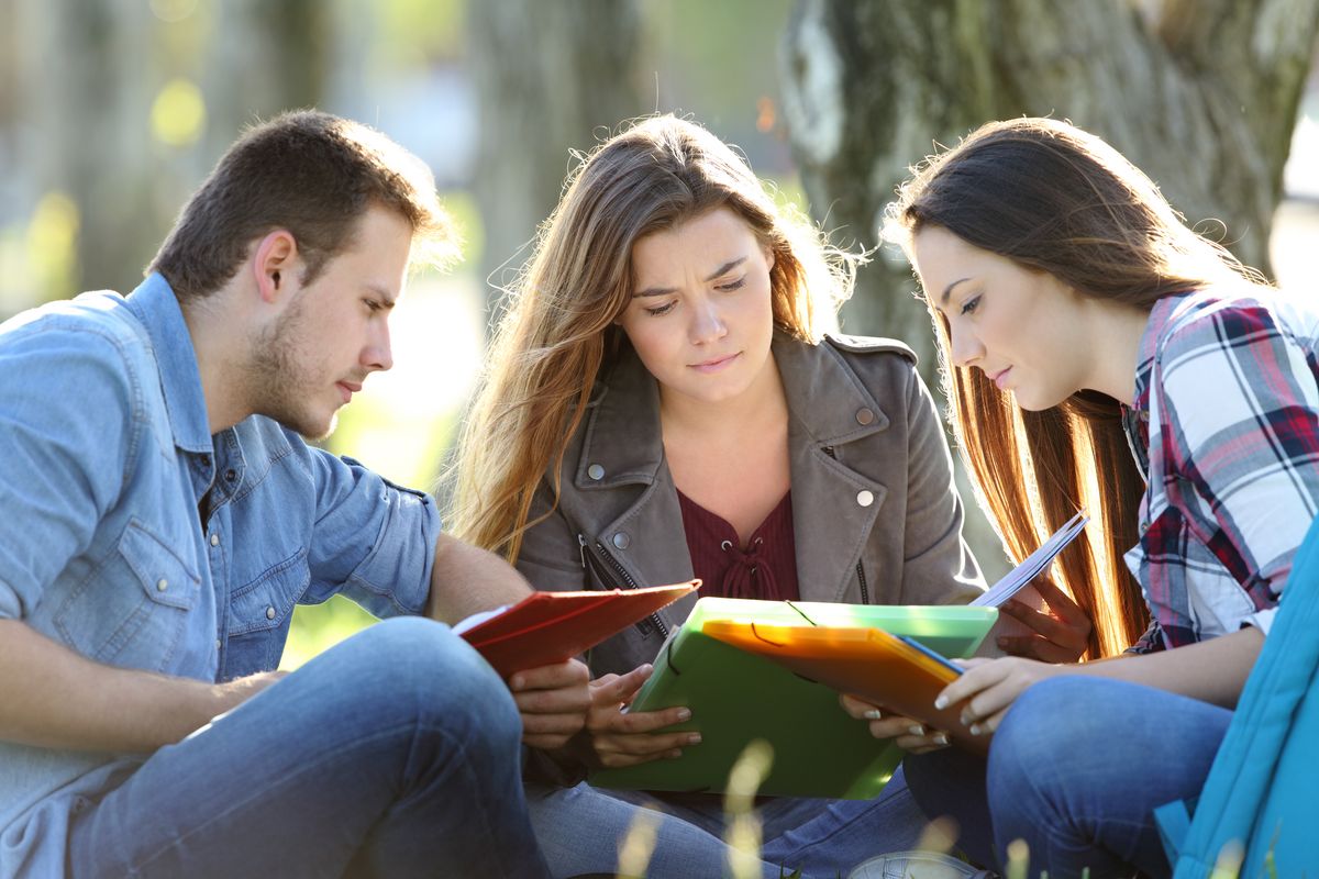 zur Vergrößerungsansicht des Bildes: Drei junge Menschen sitzen im Park und beugen sich gemeinsam über Studienunterlagen.
