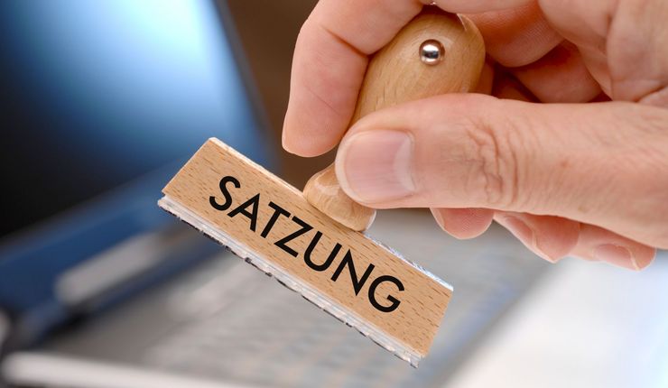 Farbfoto: Nahaufnahme einer Hand, die einen Stempel mit der Aufschrift "Satzung" in der Hand hält.