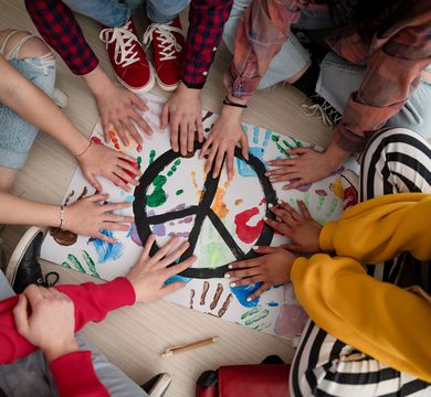 Junge Menschen sitzen im Kreis und halten ihre Hände über eine Unterlage mit farbigen Handabdrücken und einem Peace-Zeichen.