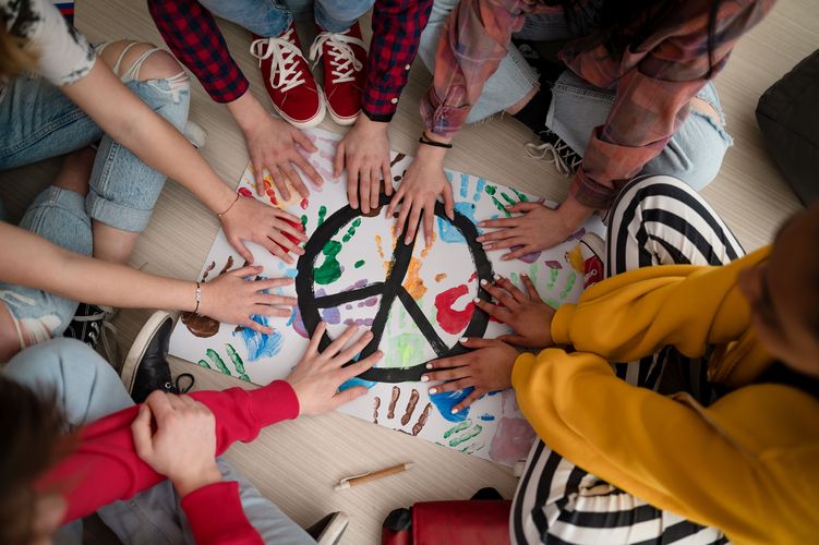 Junge Menschen sitzen im Kreis und halten ihre Hände über eine Unterlage mit farbigen Handabdrücken und einem Peace-Zeichen.