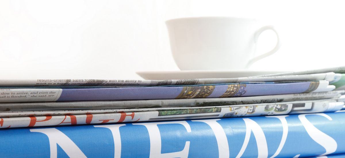 Detailfotografie: Ein Stapel Zeitungen seitlich fotografiert, auf den Zeitungen steht eine weiße Kaffeetasse, auf dem Rücken der Zeitungen ist das Wort "News" zu lesen.