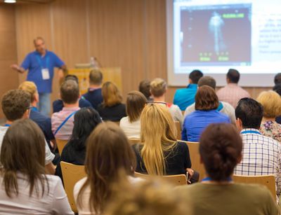 A lecturer talking in front of the audience in a lecture hall with the video presentation in the background