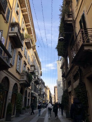 Farbfoto: Aufnahme einer engen Straße zwischen Häusern mit Balkonen und Pflanzen. Über der Straße hängen Lichterketten 