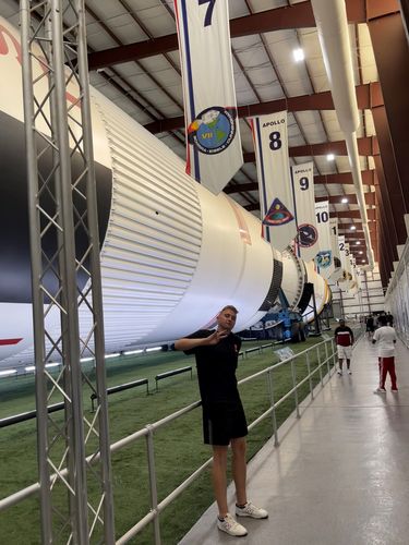 Jonas ist im NASA Museum in Houston. Er lächelt in die Kamera. Hinter ihm ist eine Rakete zu sehen, die seitlich gelagert ist.