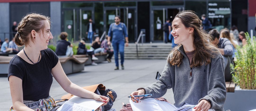 Two female students sit cross-legged on a bench in the university courtyard and talk. In the background, people walking into the lecture hall building can be seen out of focus.