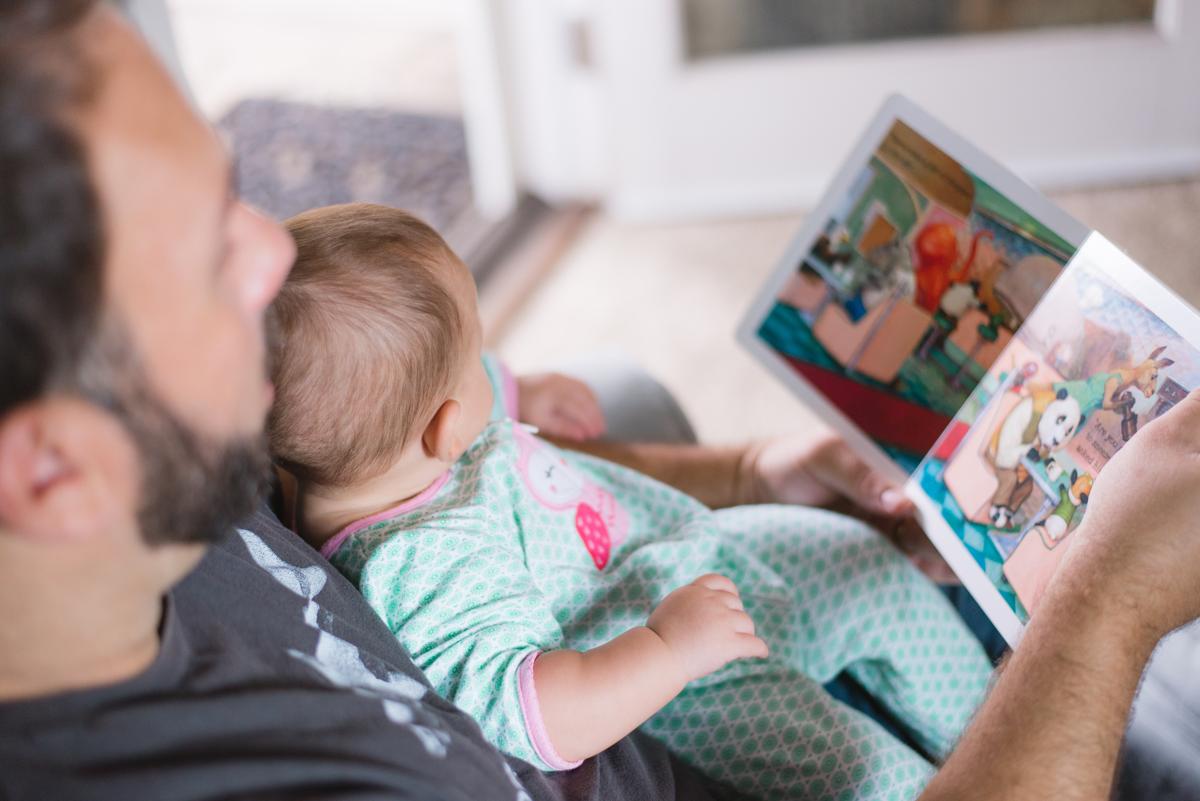 zur Vergrößerungsansicht des Bildes: Junger Mann schaut mit einem Kleinkind auf dem Schoß in ein Bilderbuch